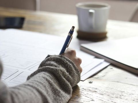 A person writing on paperwork at a wooden table beside a coffee mug